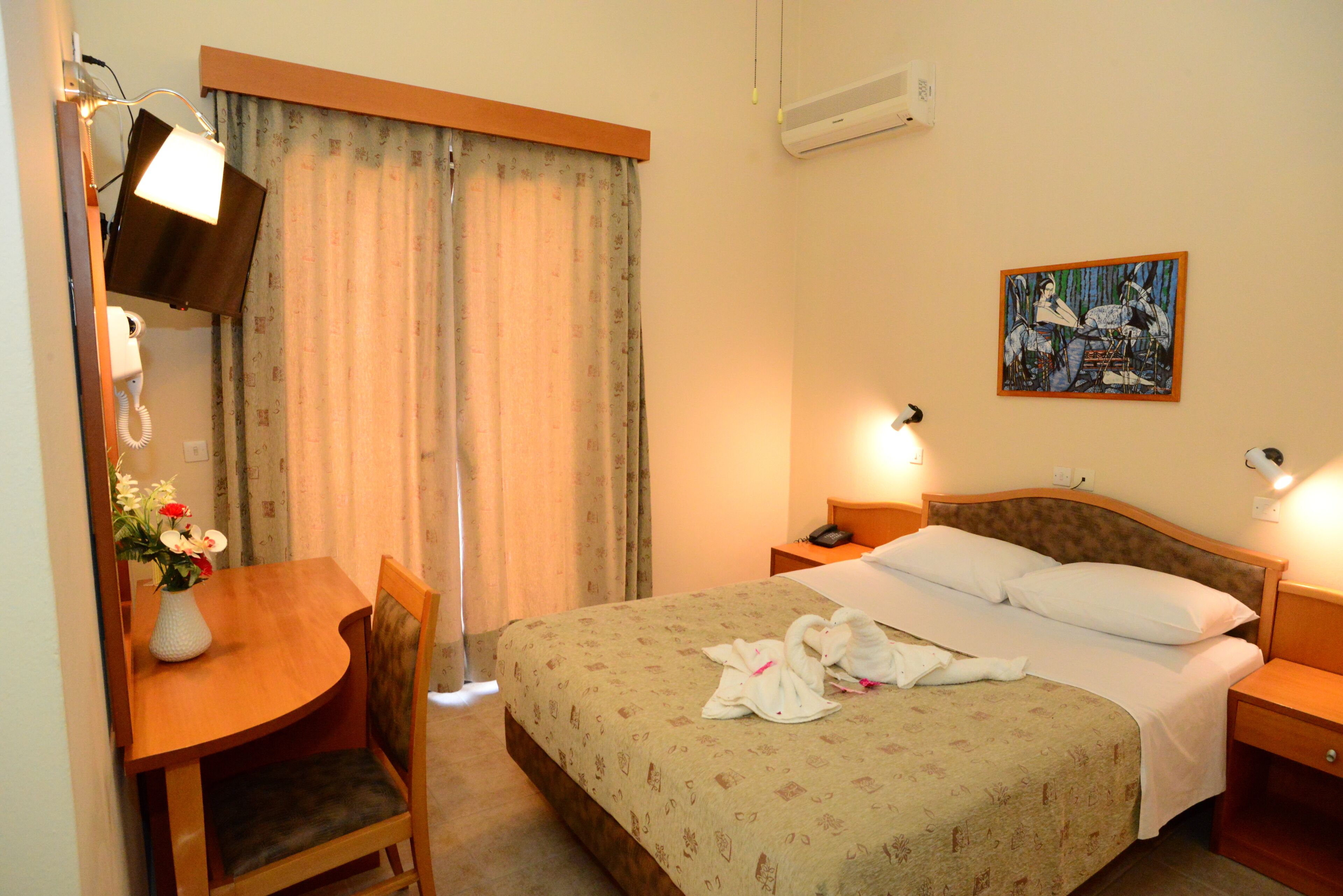 A hotel room with a bed, a desk with a lamp, a chair, a painting on the wall, and a window with curtains.