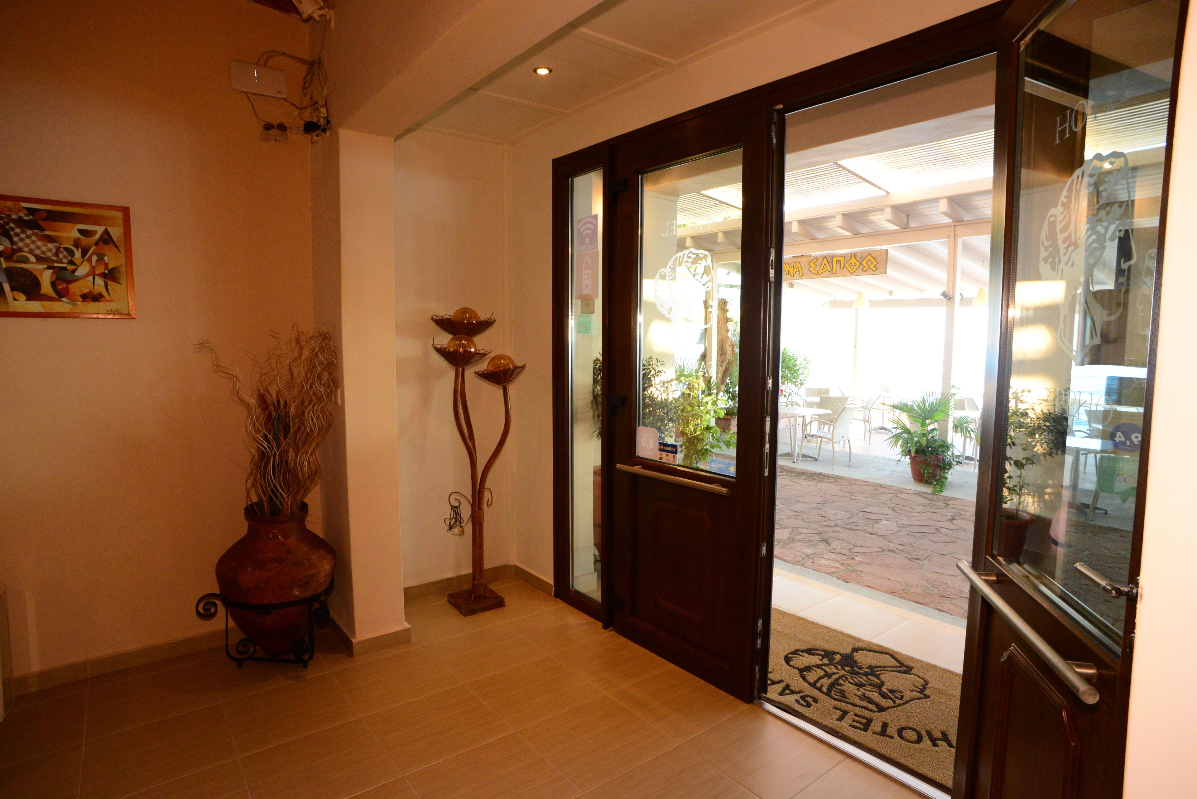 A hotel entrance with a wooden floor, a doormat with a logo, a vase with dried plants, and a painting on the wall.