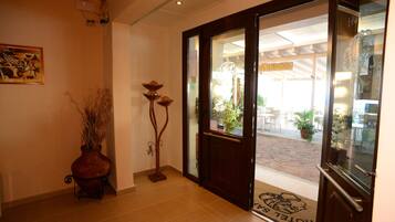 A hotel entrance with a wooden floor, a doormat with a logo, a vase with dried plants, and a painting on the wall.
