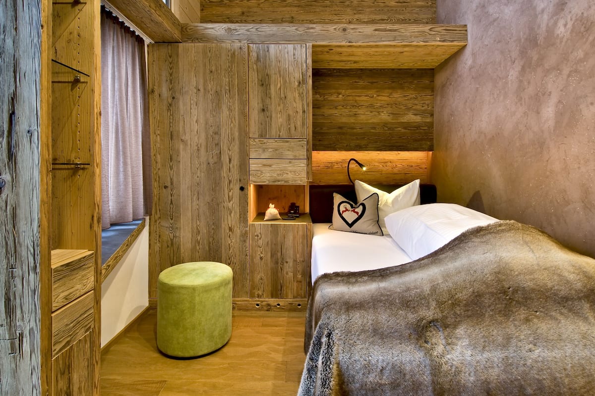 A cozy bedroom with a wooden wardrobe, a bedside table with a lamp, a green ottoman, and a bed with a heart-shaped pillow.
