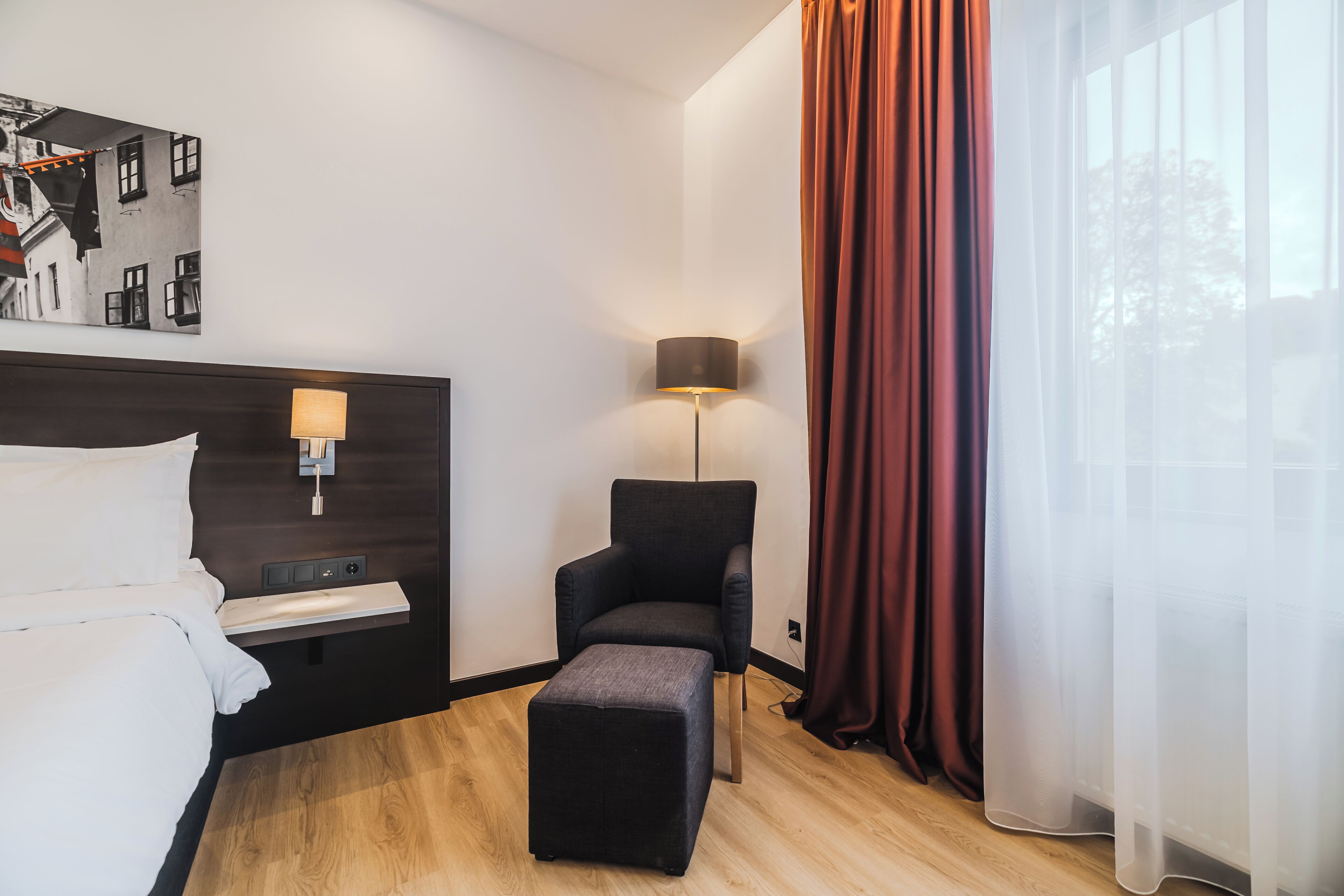 A hotel room with a bed, a desk, a chair, a lamp, and a window with curtains.