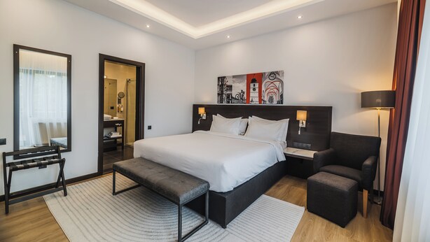 A modern hotel room with a large bed, bedside tables, a bench, a chair, and a painting on the wall. - Hotel Republique (Sibiu)
