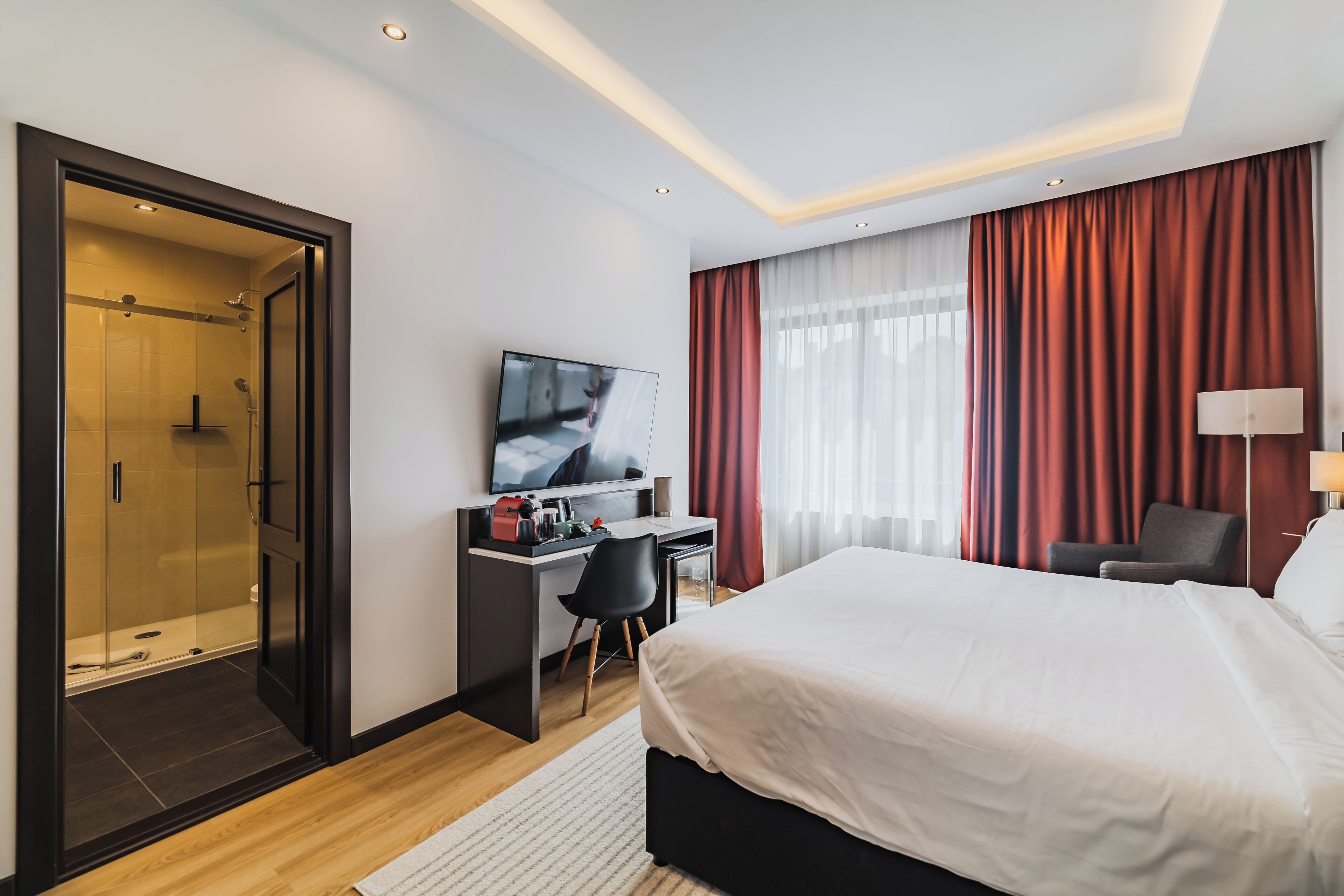 Une chambre d’hôtel moderne dotée d’un grand lit, d’un bureau avec une télévision, d’une douche et offrant une vue sur la ville grâce à de grandes fenêtres.