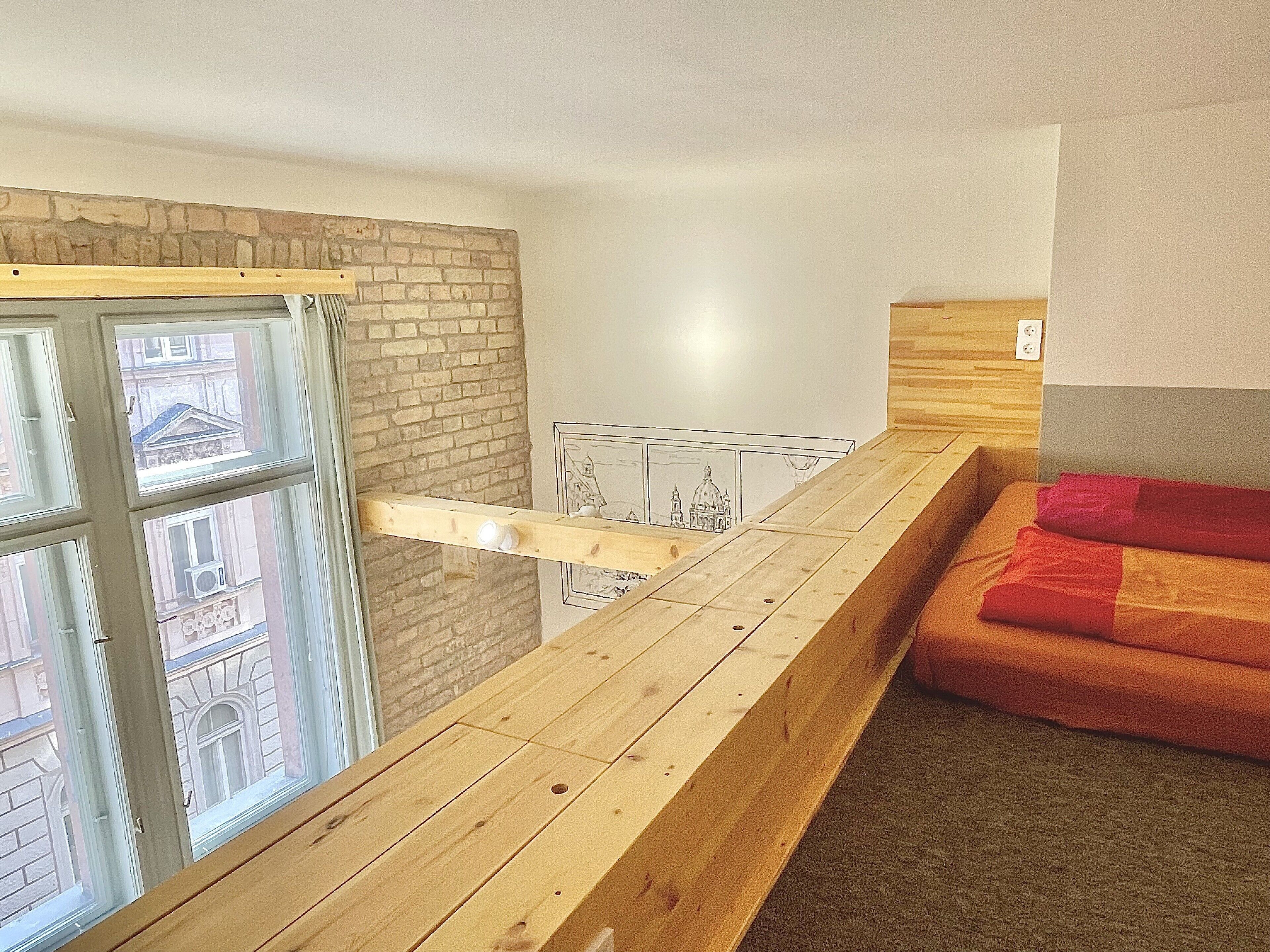A room with a wooden balcony, a red sofa, and a view of buildings through a window with a curtain.