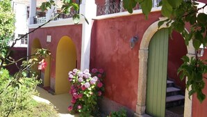 A building with a red facade, yellow arched doorways, and a green door. There are potted plants and flowers in front. - Villa de Loulia Hotel (Corfu)