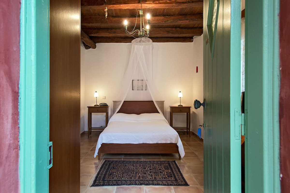 Un dormitorio con techo de madera, una cama con dosel blanco, dos mesitas de noche con lámparas y una alfombra estampada.