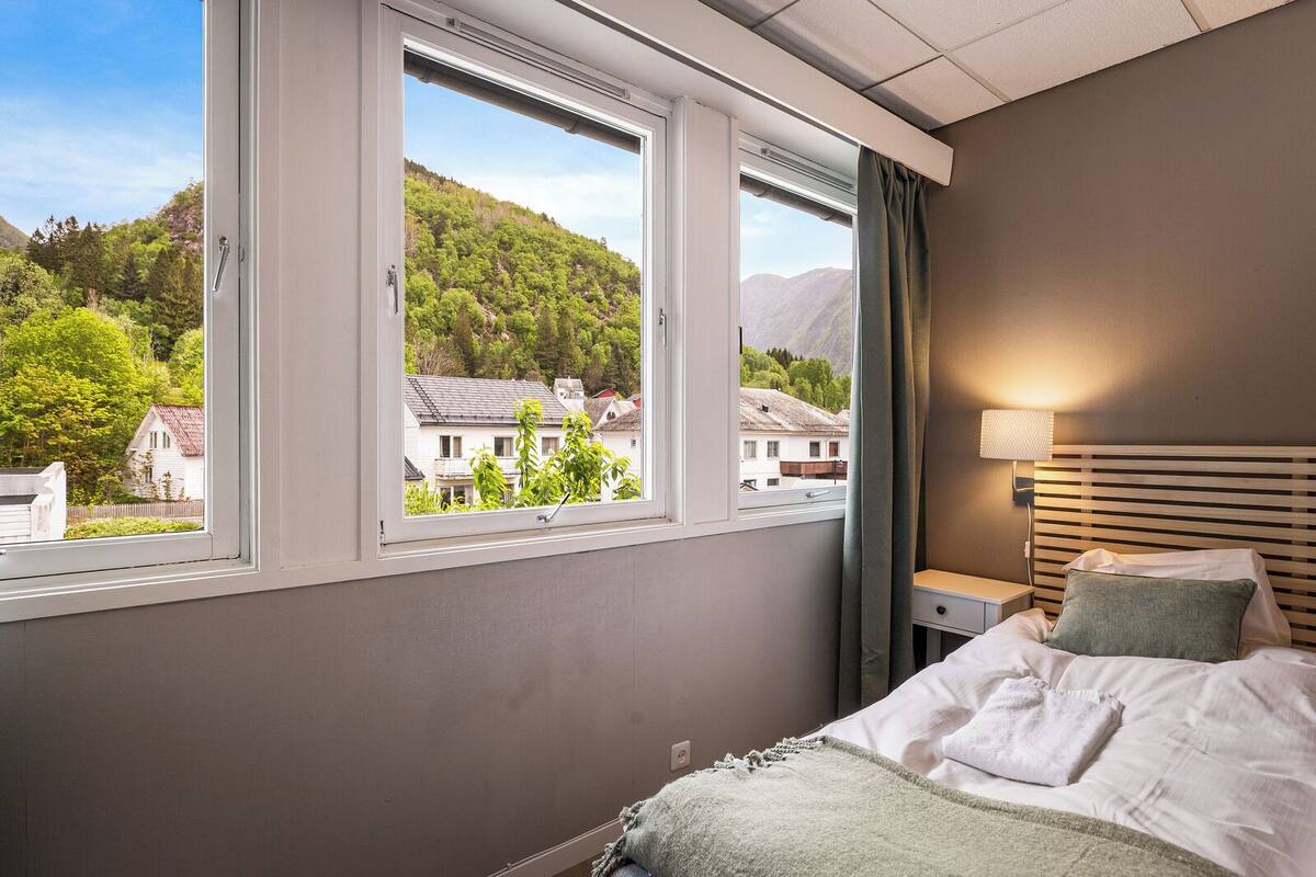A bedroom with a bed, a bedside table, a lamp, and a window with a view of houses and trees.
