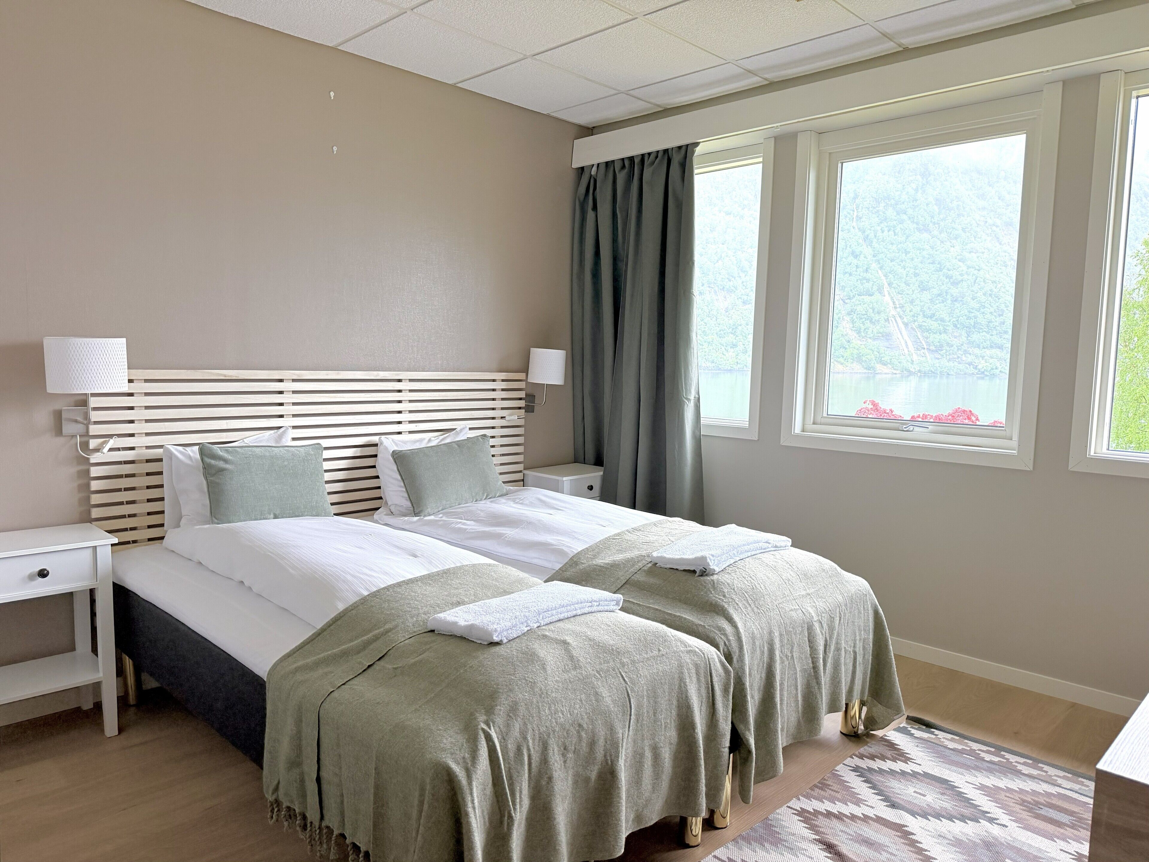 A double bed with a wooden headboard, two bedside tables, a window with curtains, and a view of greenery outside.