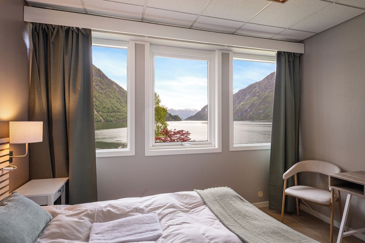 A bedroom with a bed, a desk, a chair, and a window with a view of mountains and a lake.