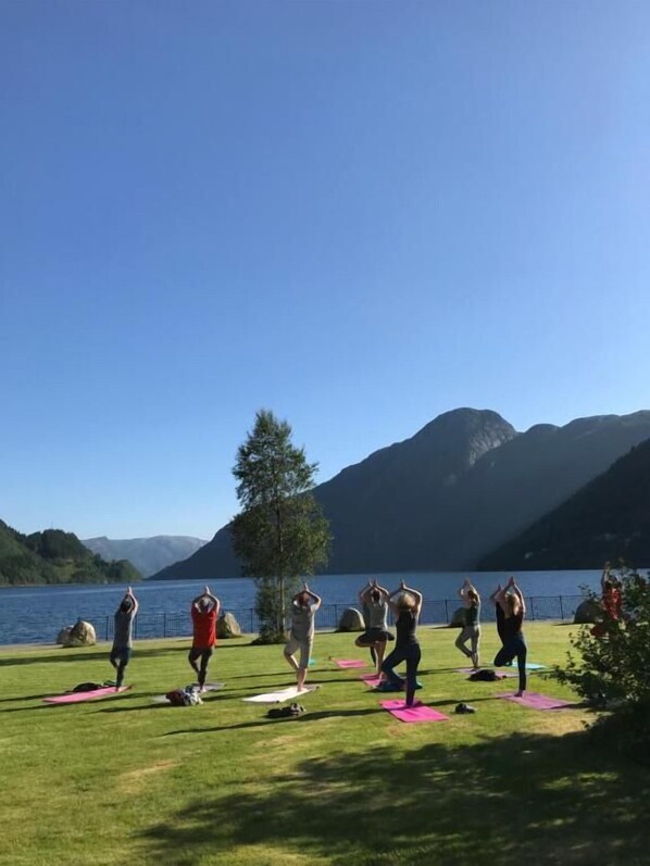 Un grupo de personas practicando yoga sobre esterillas junto a un lago con montañas al fondo.