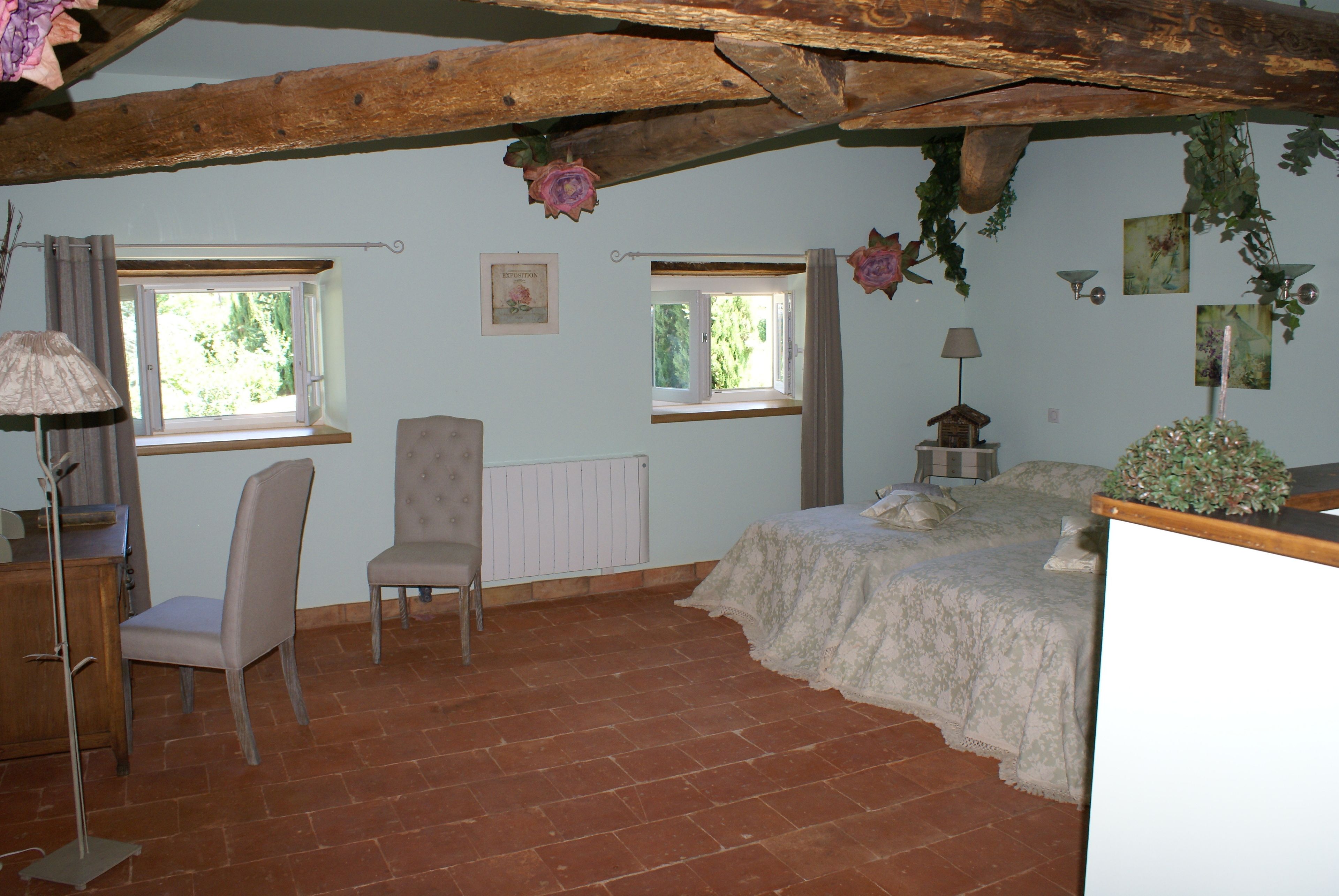 A bedroom with a rustic wooden ceiling, two windows with curtains, a bed with a floral bedspread, a chair, a desk with a lamp, and a radiator.