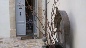 A stone-paved entrance with a blue door, a stone wall, and a water feature with a stone basin and branches. - Maison Bersane (Le Poujol-sur-Orb)