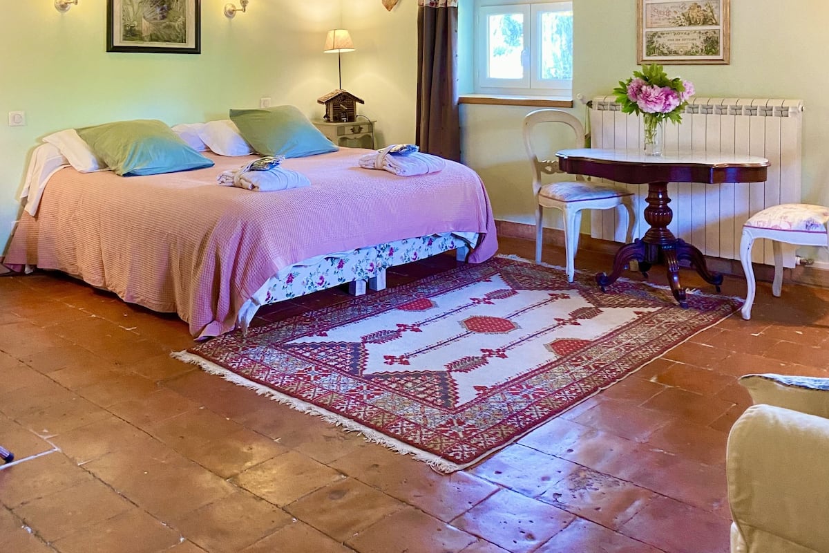 Un dormitorio rústico con vigas de madera a la vista, una cama con colcha estampada, una mesita con un florero y un cuadro en la pared.