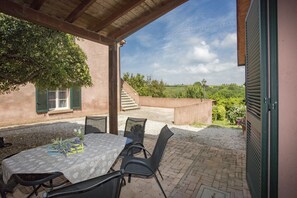 An outdoor patio with a table and chairs, a covered area, and a view of a garden and buildings in the background.
