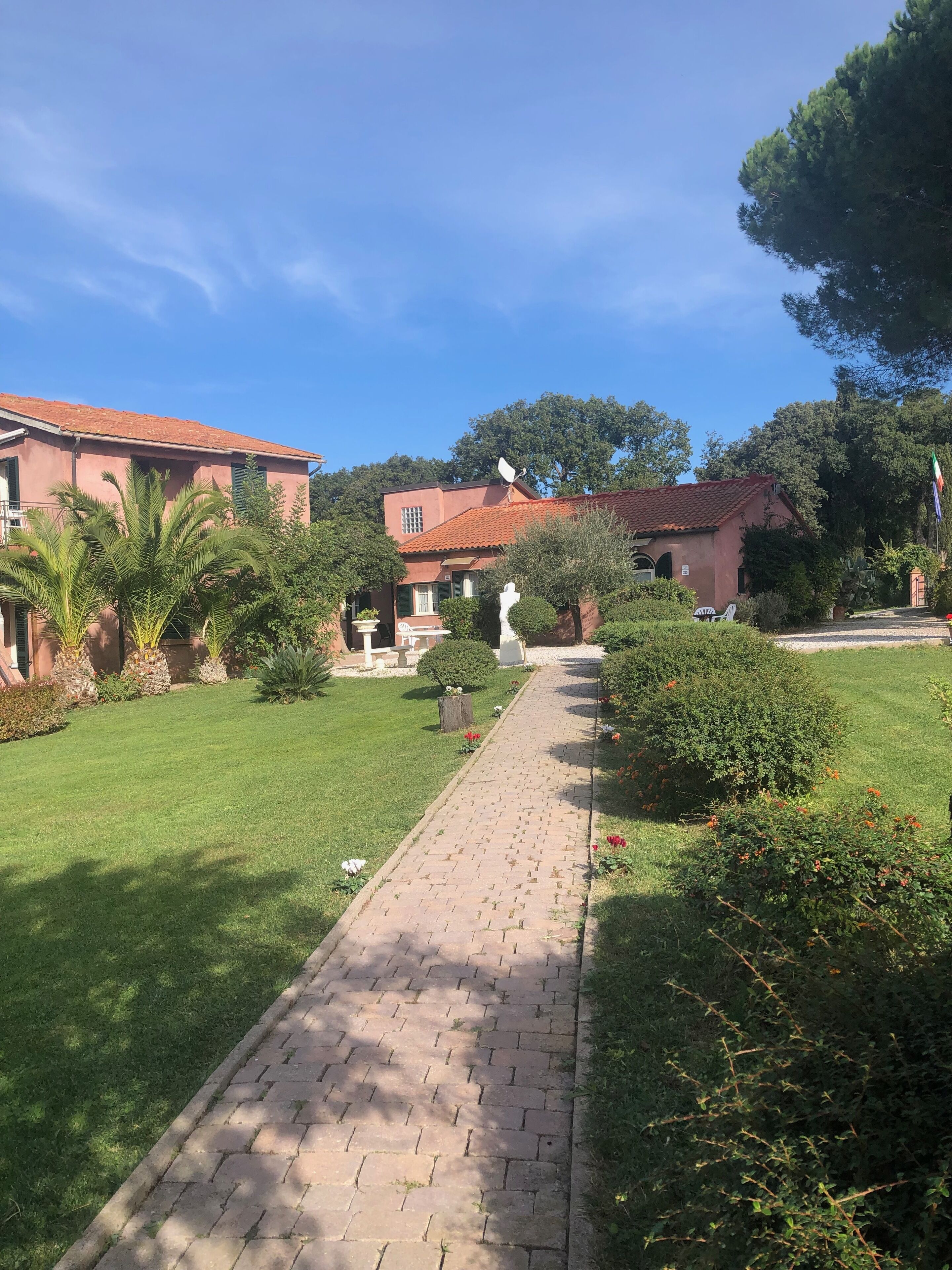 Un vialetto lastricato conduce a un edificio con tetto in tegole rosse, immerso nel verde e sotto un cielo azzurro intenso.