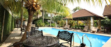 A pool area with a table and chairs, surrounded by palm trees and lounge chairs.
