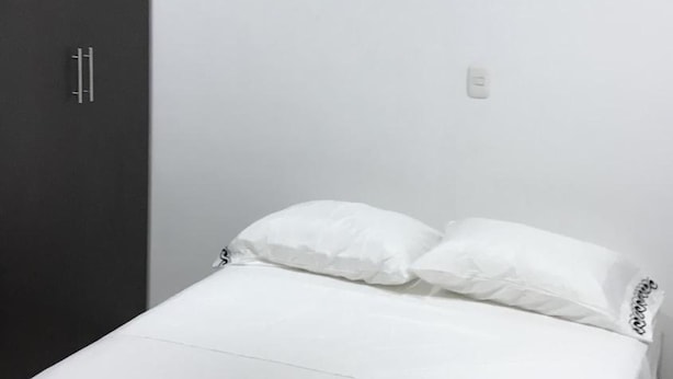A bedroom with a bed, white bedding, a bedside table, a wardrobe, a ceiling fan, and an air conditioning unit. - Barcelona Hotel (Montería)