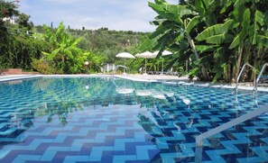 A swimming pool with a blue and white tiled floor, surrounded by lush greenery and white umbrellas. - Yanna Studios & Apartments (Zakynthos)