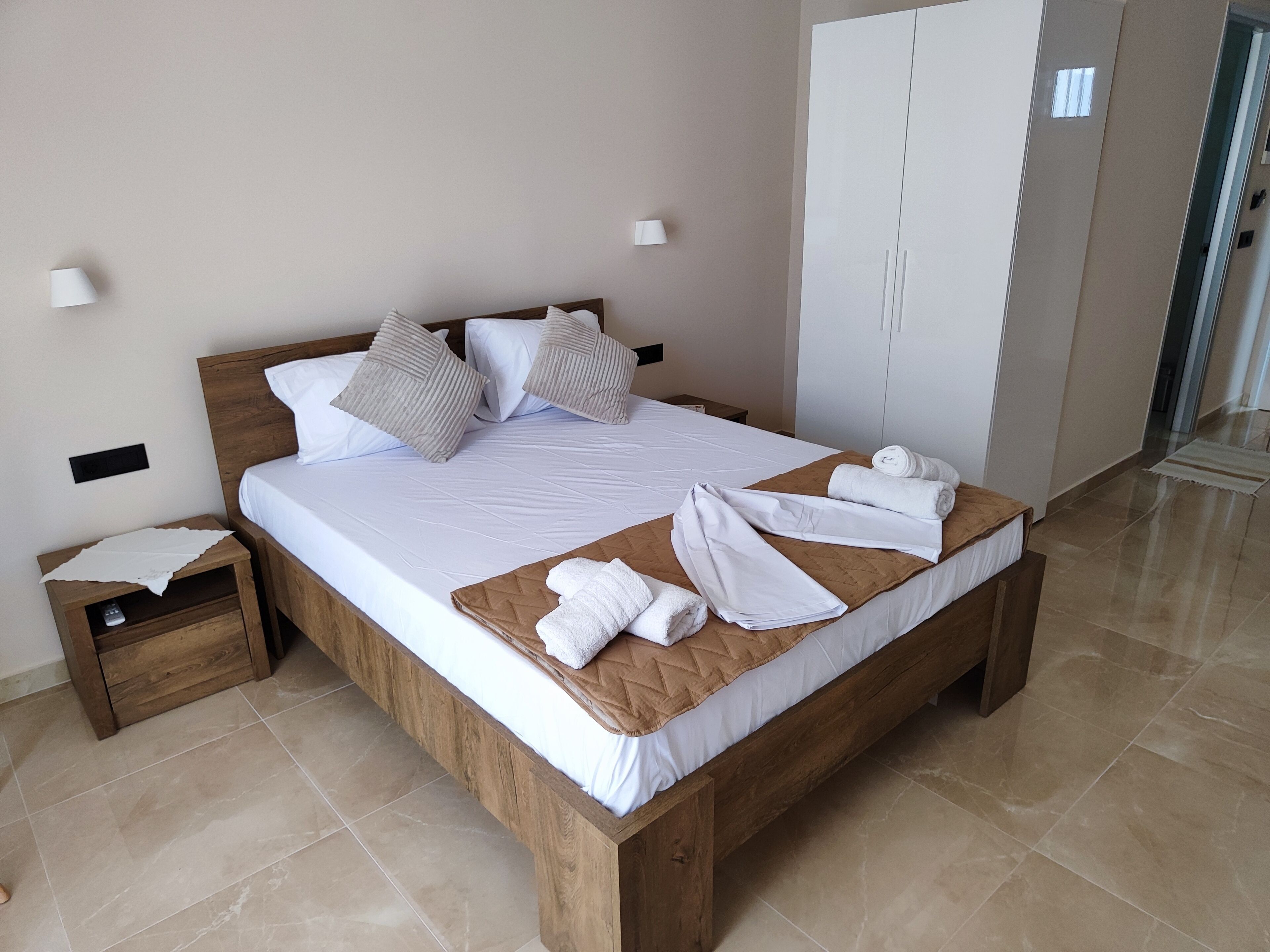A neatly made bed with white linens and pillows, a wooden nightstand with a remote control, and a pair of towels on the bed.