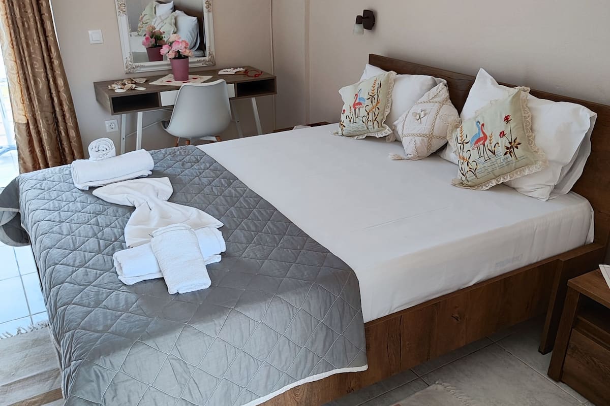 A neatly made bed with a grey quilt, white pillows, and a wooden headboard. A desk with a mirror and a chair is positioned against the wall.