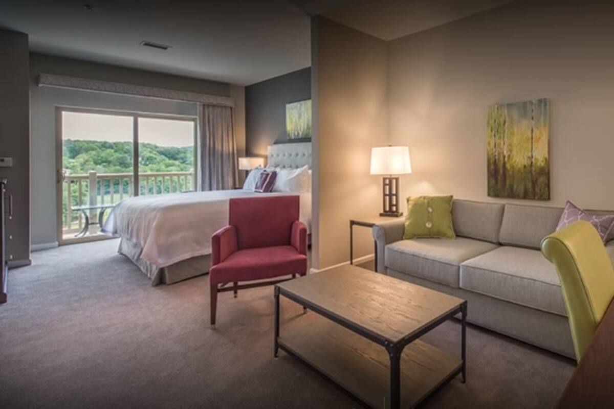 A hotel room with a bed, a sofa, a red armchair, a wooden coffee table, and a desk with a telephone.