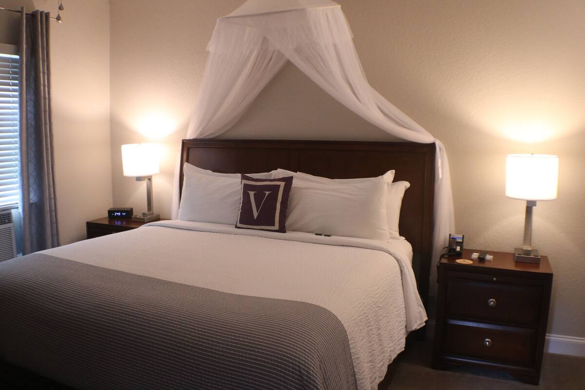 A hotel room with a canopy bed, two bedside lamps, a nightstand with a clock, and a bedside table with a lamp.
