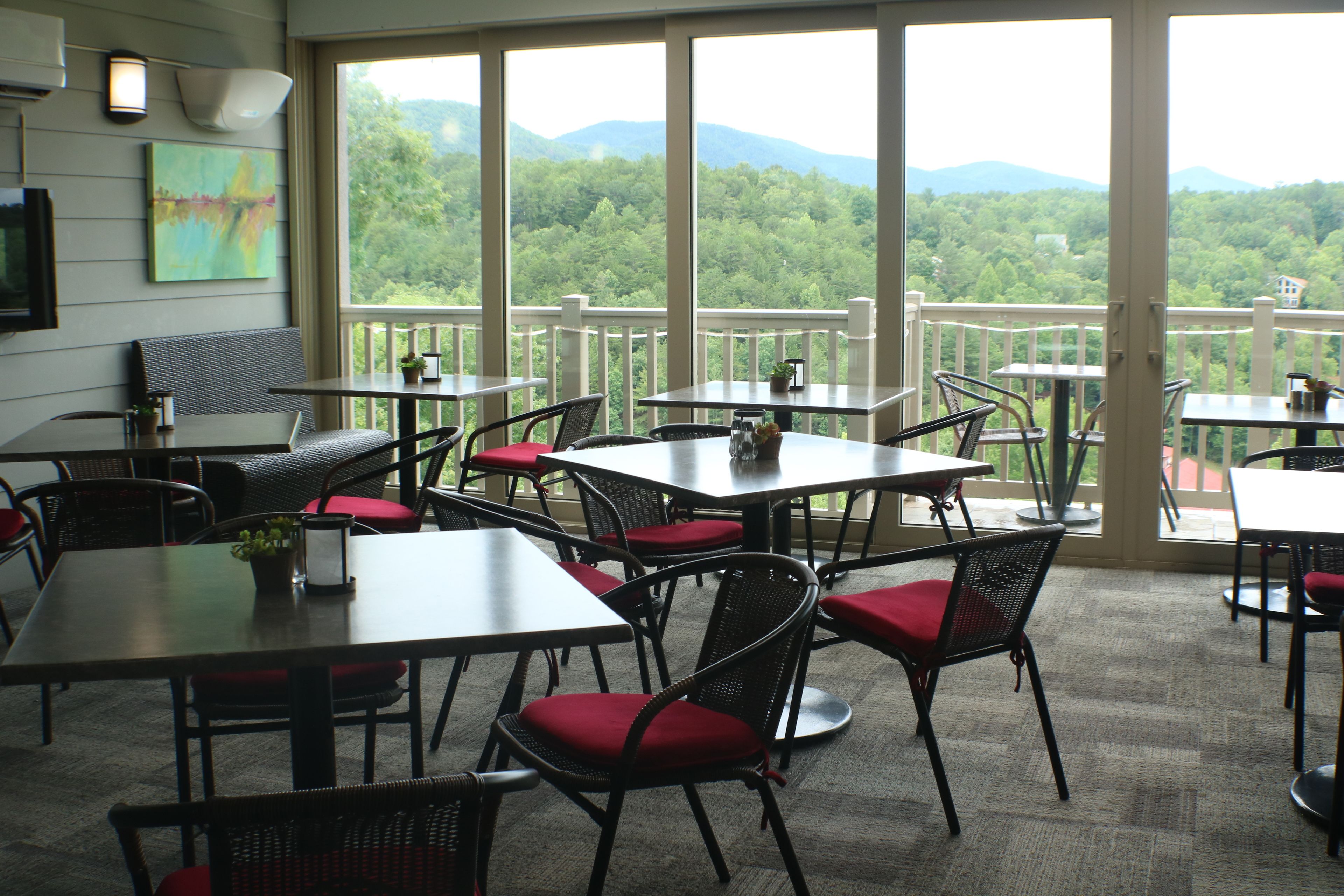 A restaurant with multiple tables and chairs, large windows with a view of a forested mountain range.