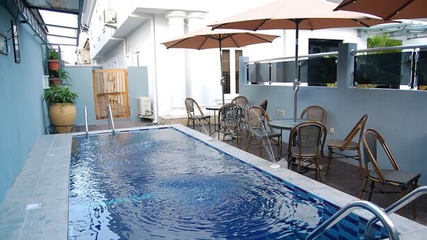 Una piscina con borde de azulejos, rodeada de mobiliario exterior con sombrillas y una planta en maceta a la izquierda.