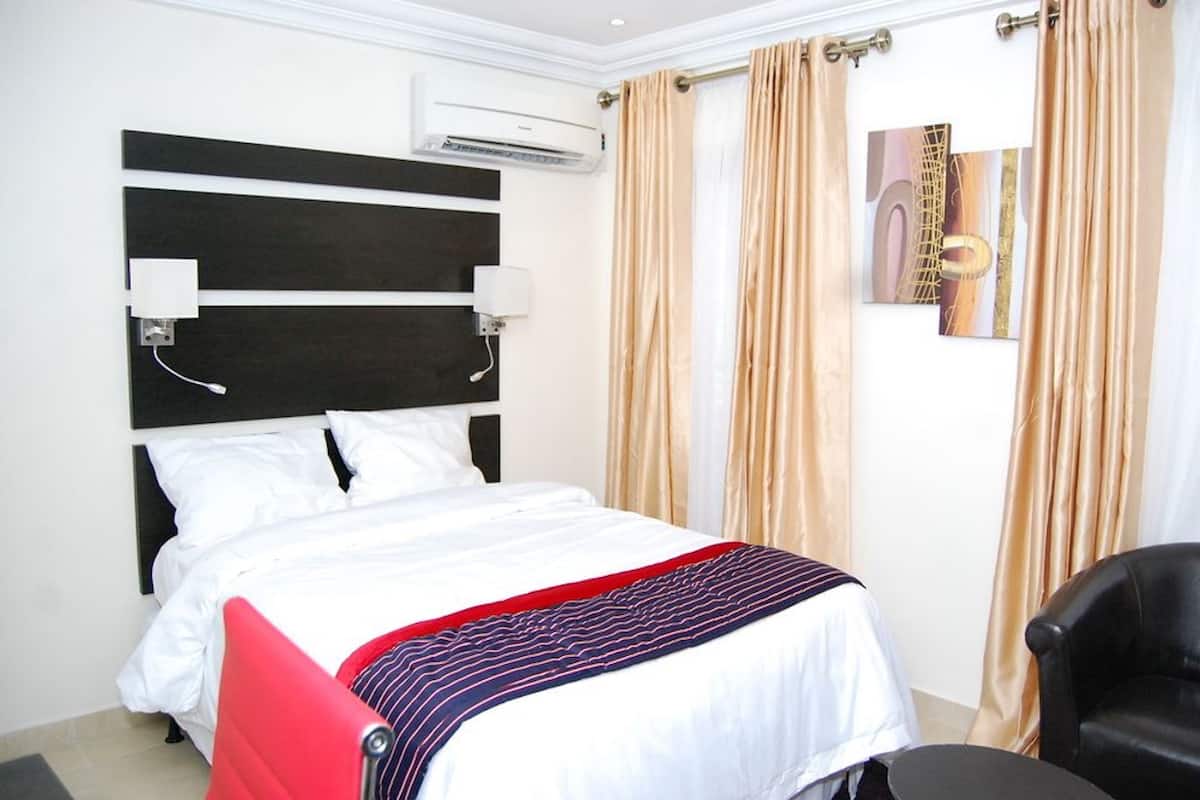 A hotel room with a bed, a red chair, a black chair, beige curtains, and a wall-mounted air conditioner.