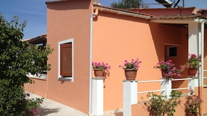 A building with a flat roof, a window with wooden shutters, and several potted plants with pink flowers. - Ionian Breeze Apartments (Corfu)