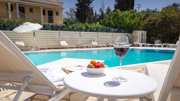 A poolside setting with a table, a bowl of fruit, and a glass of red wine. - Ionian Breeze Apartments (Corfu)