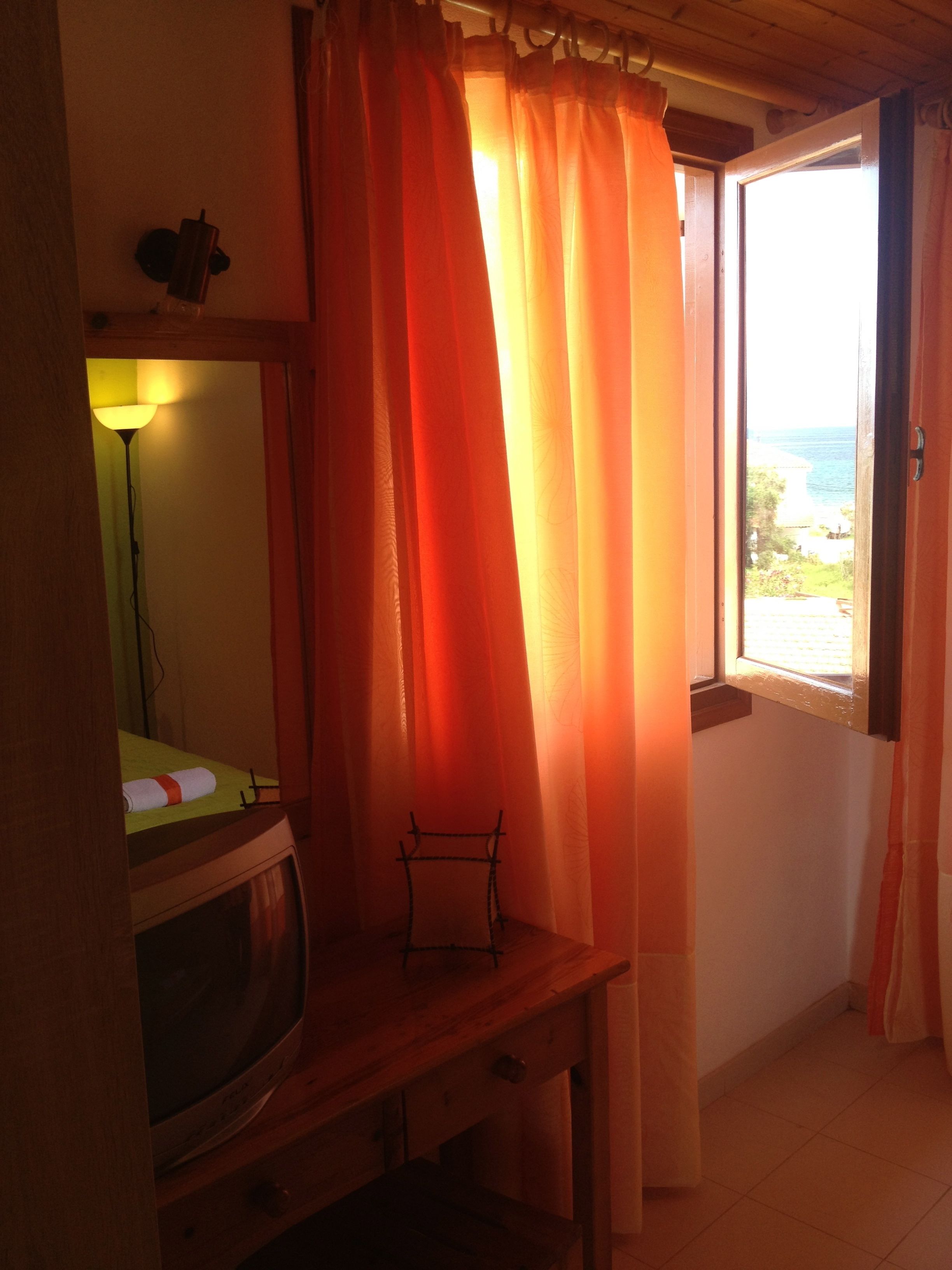 Ein Zimmer mit Schreibtisch aus Holz, einem Fernseher und Meerblick durch ein Fenster mit orangefarbenen Vorhängen.