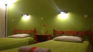 A room with two single beds, each with a wooden headboard and green bedding. There are two bedside tables, one with a vase of flowers. The walls are painted green, and there are two wall-mounted lights. - Ionian Breeze Apartments (Corfu)