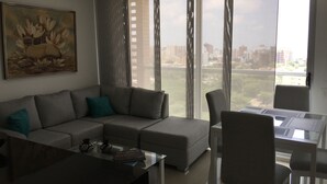A modern living room with a grey sofa, a glass coffee table, and a dining table with chairs. There is a large window with curtains, and a painting of flowers on the wall. - Apartamento Ejecutivo Ancora (Barranquilla)