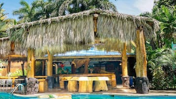A pool area with a thatched roof structure, wooden seating, and a bar area.