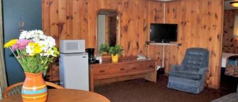 A room with wooden paneling, a television, a small refrigerator, a sofa, and a dining table with a vase of flowers.
