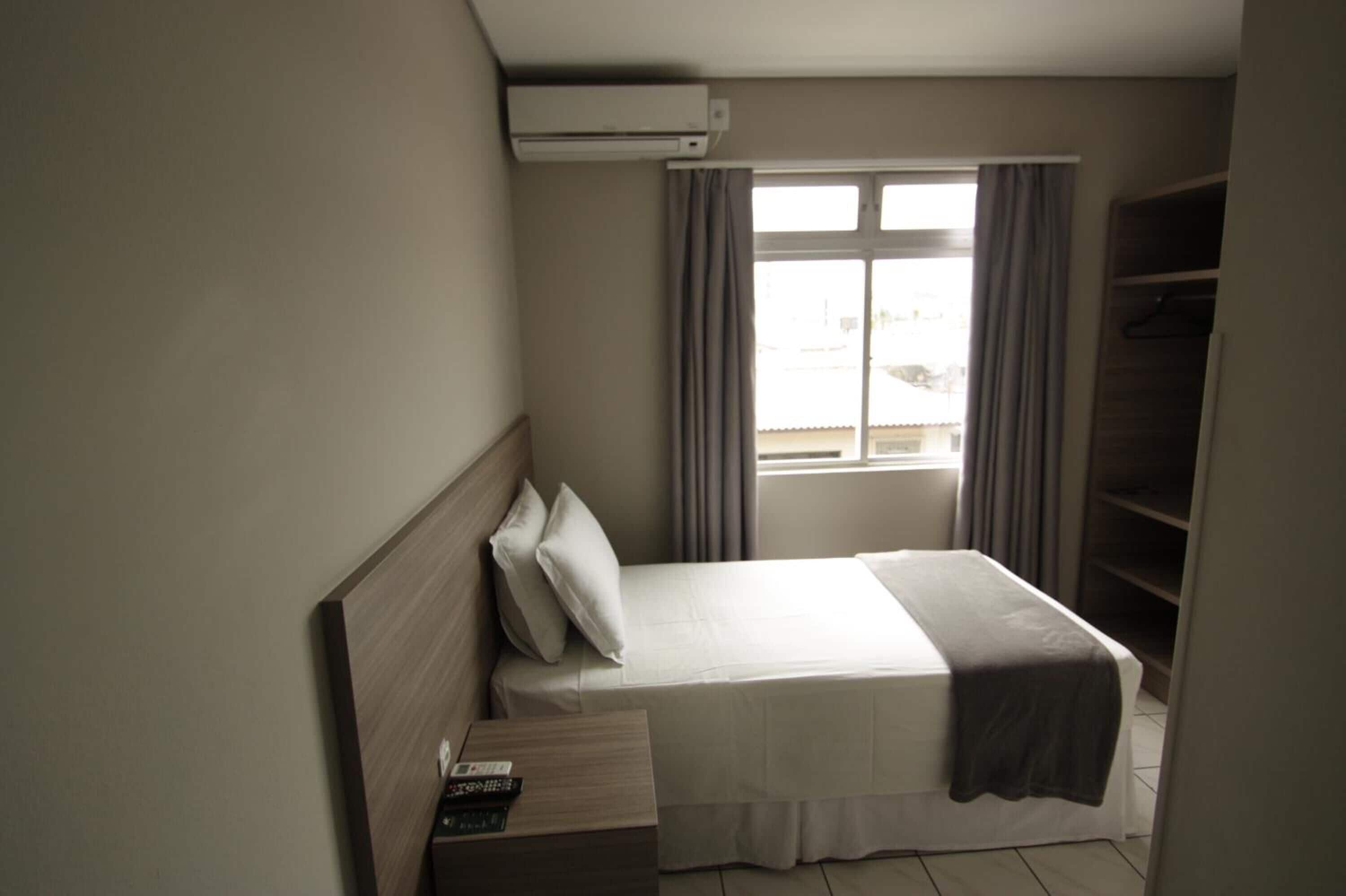 A small, neatly arranged bedroom with a single bed, a bedside table, a window with curtains, and a built-in wardrobe.