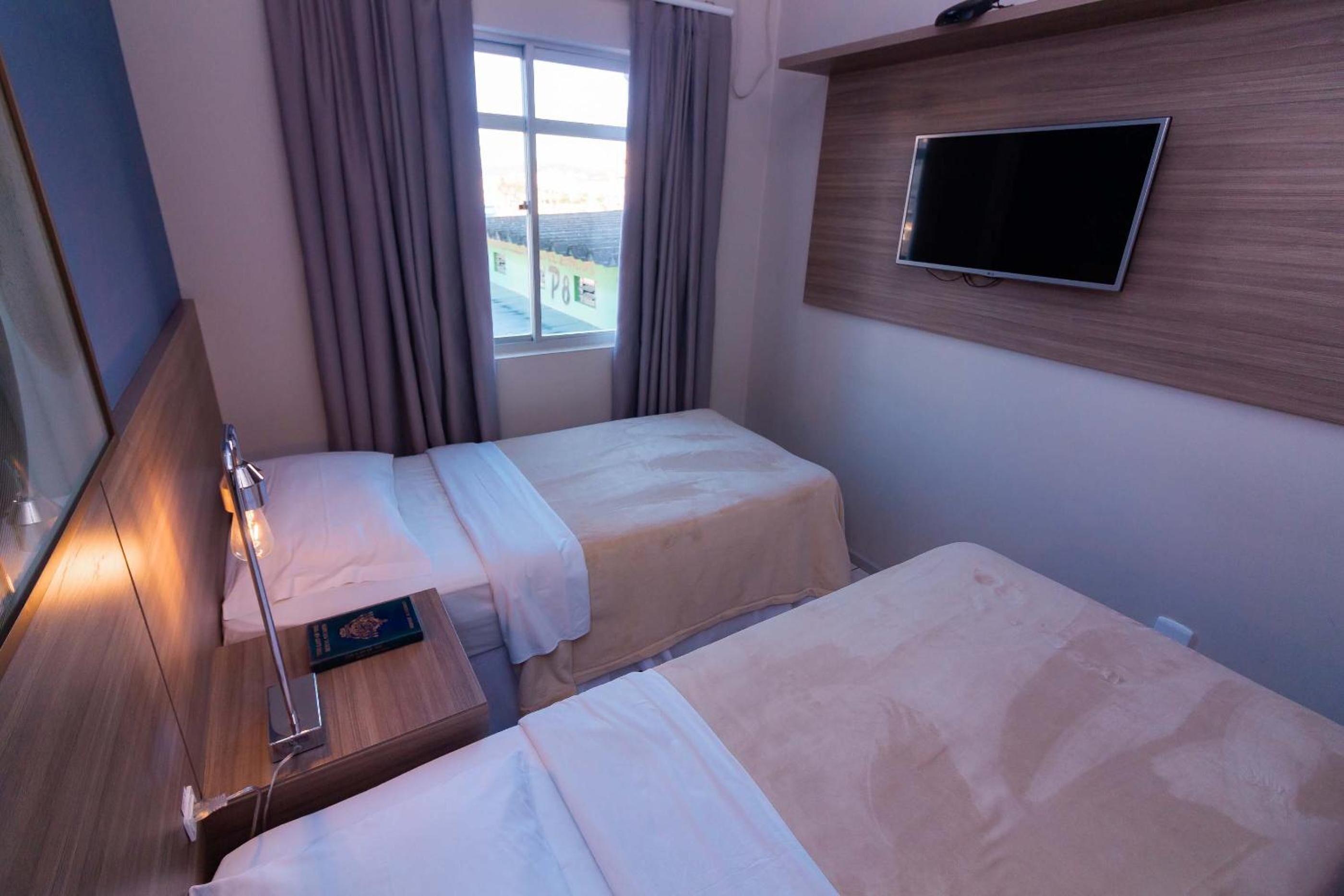 A hotel room with two beds, a TV mounted on the wall, a window with curtains, and a bedside table with a lamp.