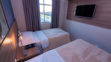 A hotel room with two beds, a TV mounted on the wall, a window with curtains, and a bedside table with a lamp.