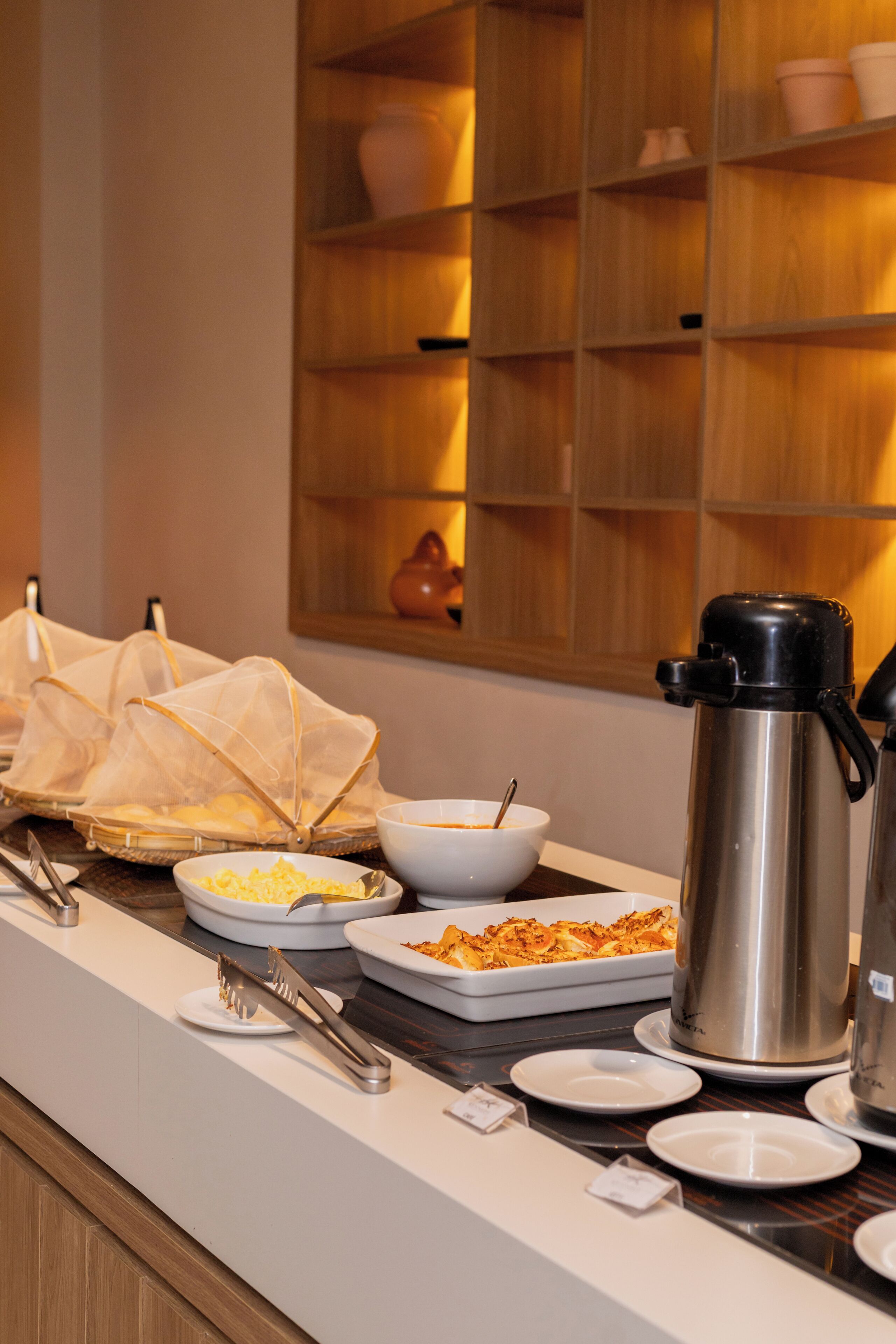 A buffet setup with various dishes, a coffee maker, and a shelf with cups and bowls.