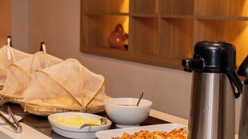 A buffet setup with various dishes, a coffee maker, and a shelf with cups and bowls.