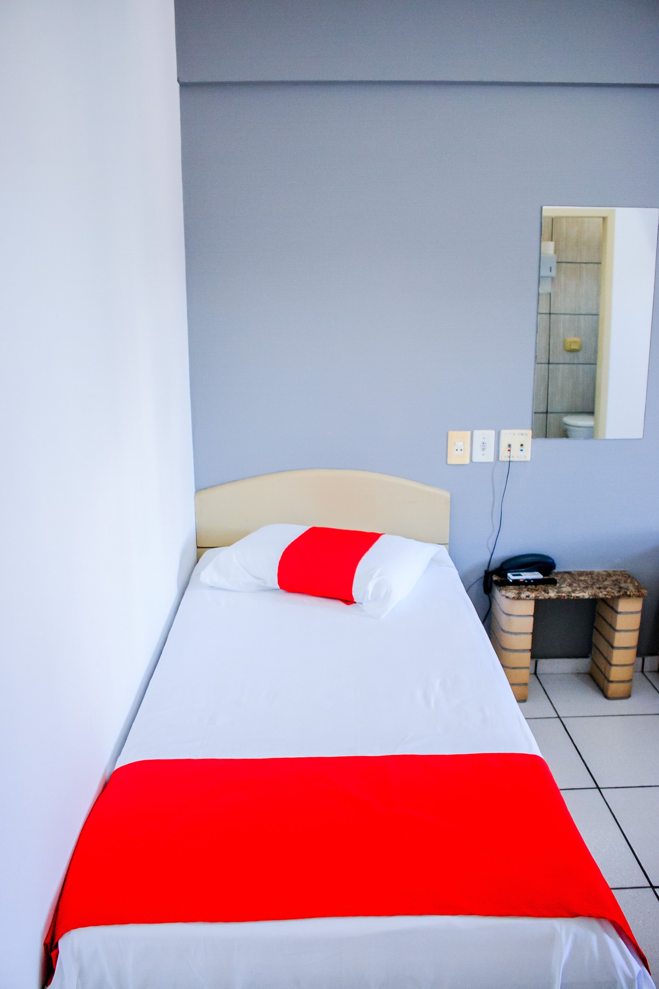 Una cama individual con una colcha blanca y roja, un cabecero color beige y una mesita de noche pequeña con un teléfono y un enchufe.