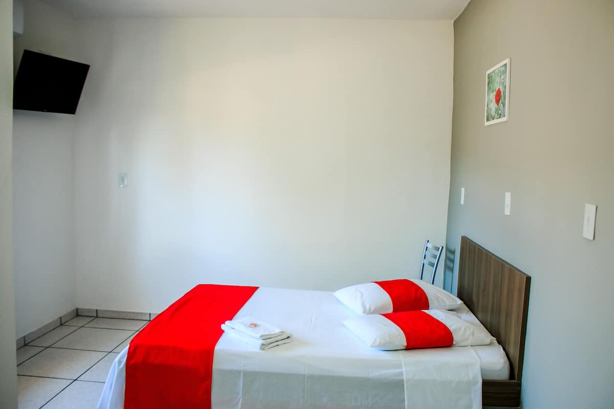 A neatly made bed with white and red bedding, a flat-screen TV mounted on the wall, and a small picture frame on the wall.