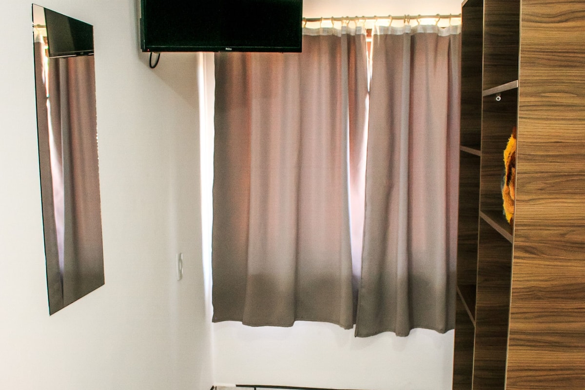 A room with a bed, a TV mounted on the wall, a wardrobe, and a window with curtains.