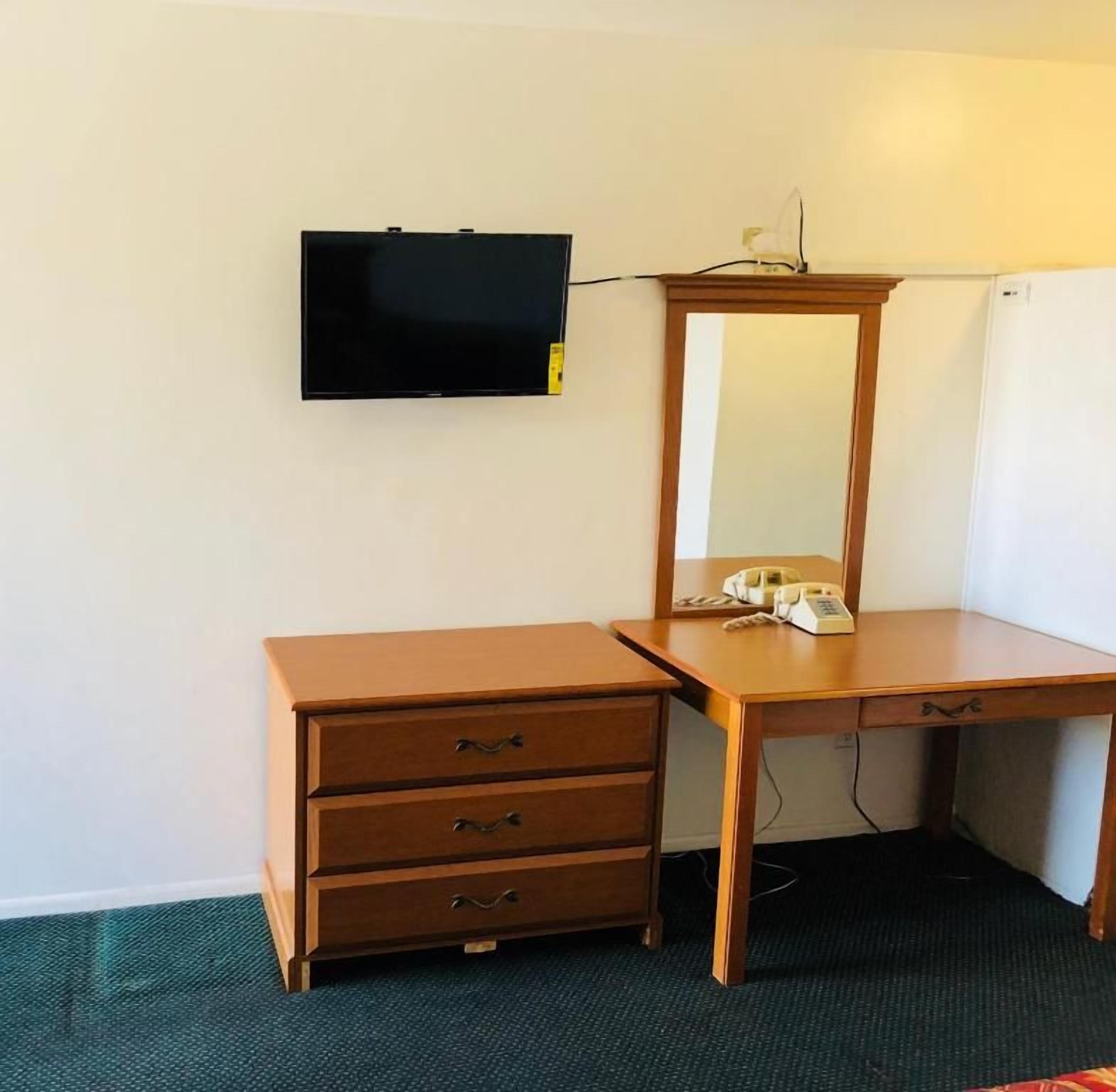 Una habitación con un escritorio de madera, un espejo, un televisor montado en la pared y un teléfono sobre el escritorio.