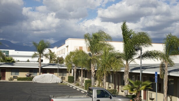 A parking lot with a silver pickup truck, a covered vehicle, and a building with multiple rooms and palm trees. - Sands Motel (Ontario)