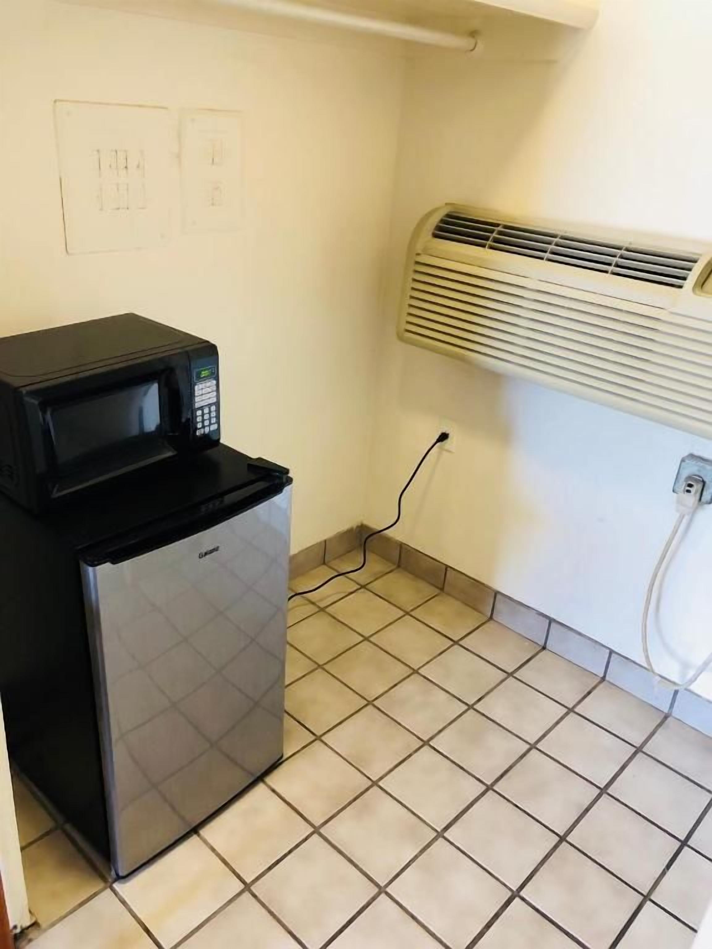 Una kitchenette compacta con microondas y refrigerador, piso de baldosas y un equipo de aire acondicionado en la pared.