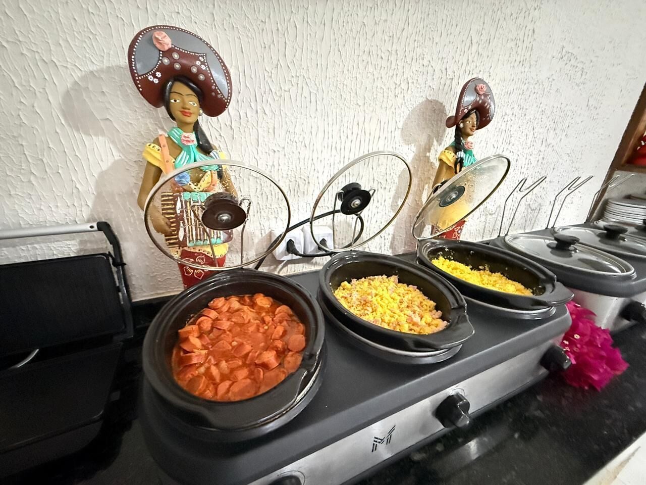 Disposição de buffet com diversos alimentos em recipientes pretos, uma figura decorativa e uma parede texturizada branca ao fundo.