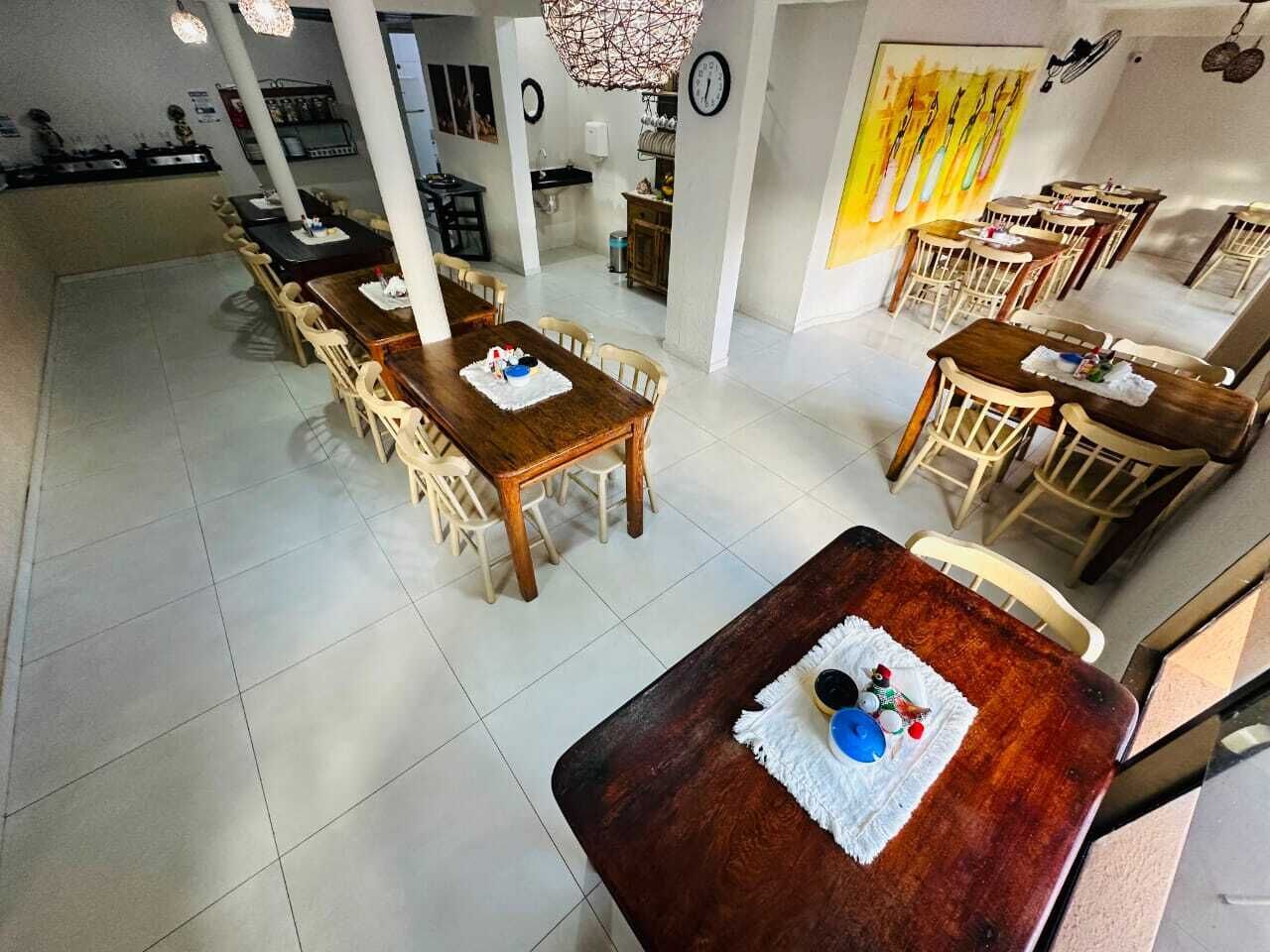 Uma ampla área de jantar com várias mesas e cadeiras de madeira, uma cozinha ao fundo e um quadro grande na parede.