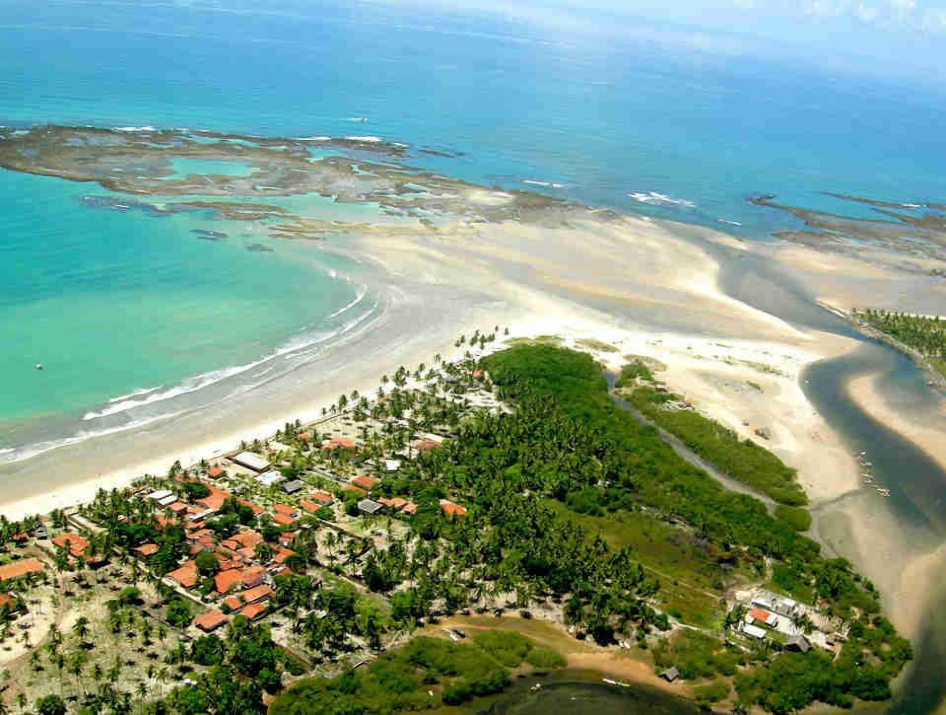 Vista aérea de una zona costera con playa, un pequeño poblado con edificios de techo rojo y abundante vegetación.
